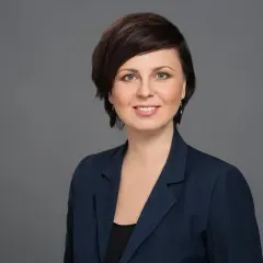 Portret uśmiechniętej kobiety w granatowej marynarce na szarym tle.
