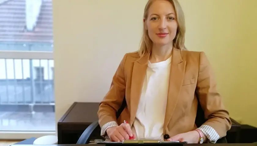 Kobieta w beżowej marynarce siedzi przy biurku i pisze.