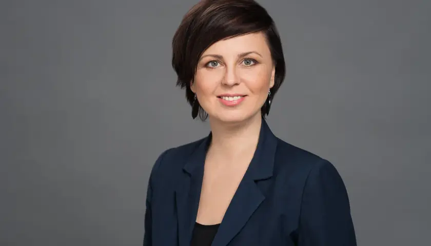 Portret uśmiechniętej kobiety w granatowej marynarce na szarym tle.